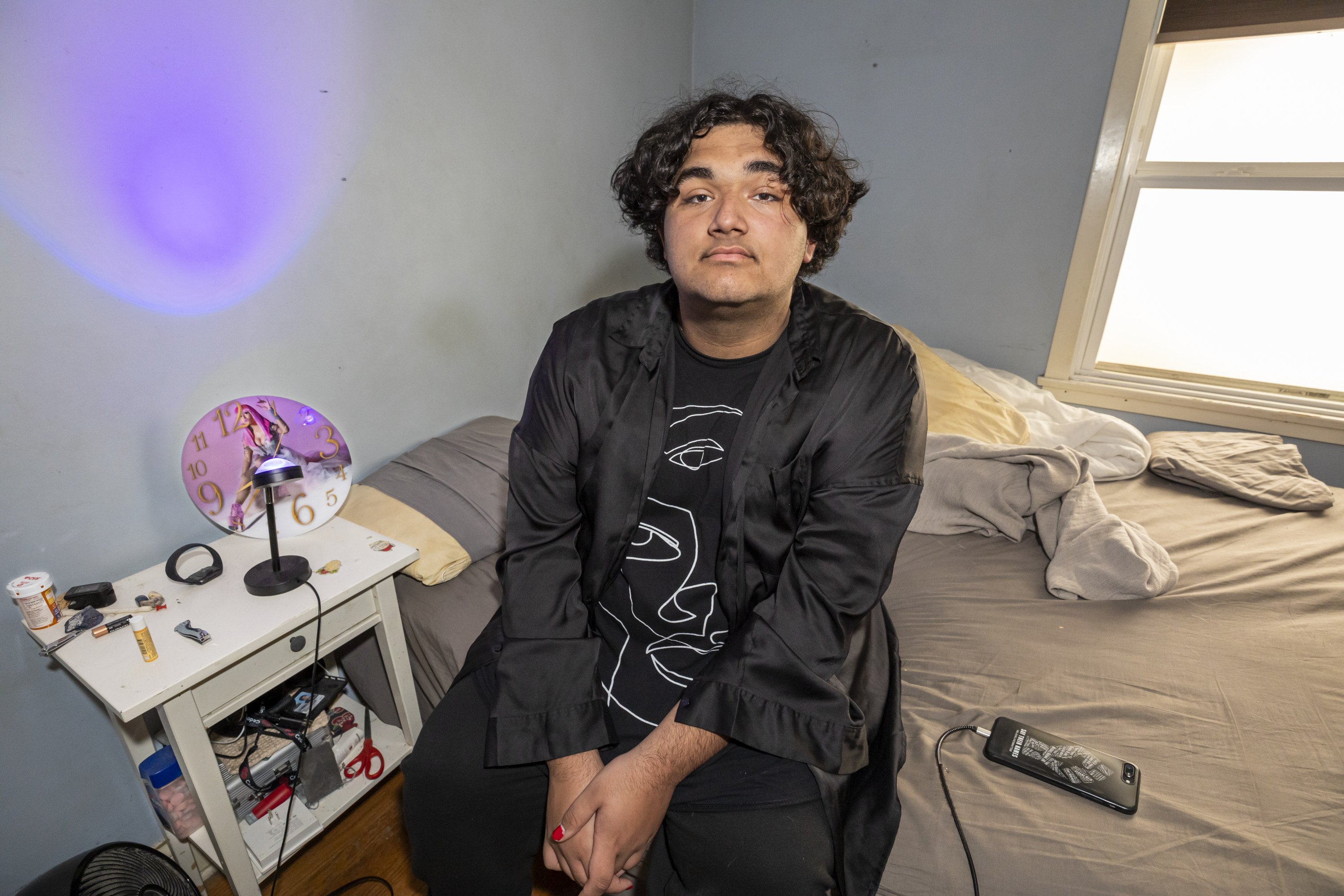 Ein junger Mann sitzt auf der Bettkante seines Zimmers und blickt in die Kamera; neben ihm auf der Decke liegt ein Smartphone am Ladekabel.