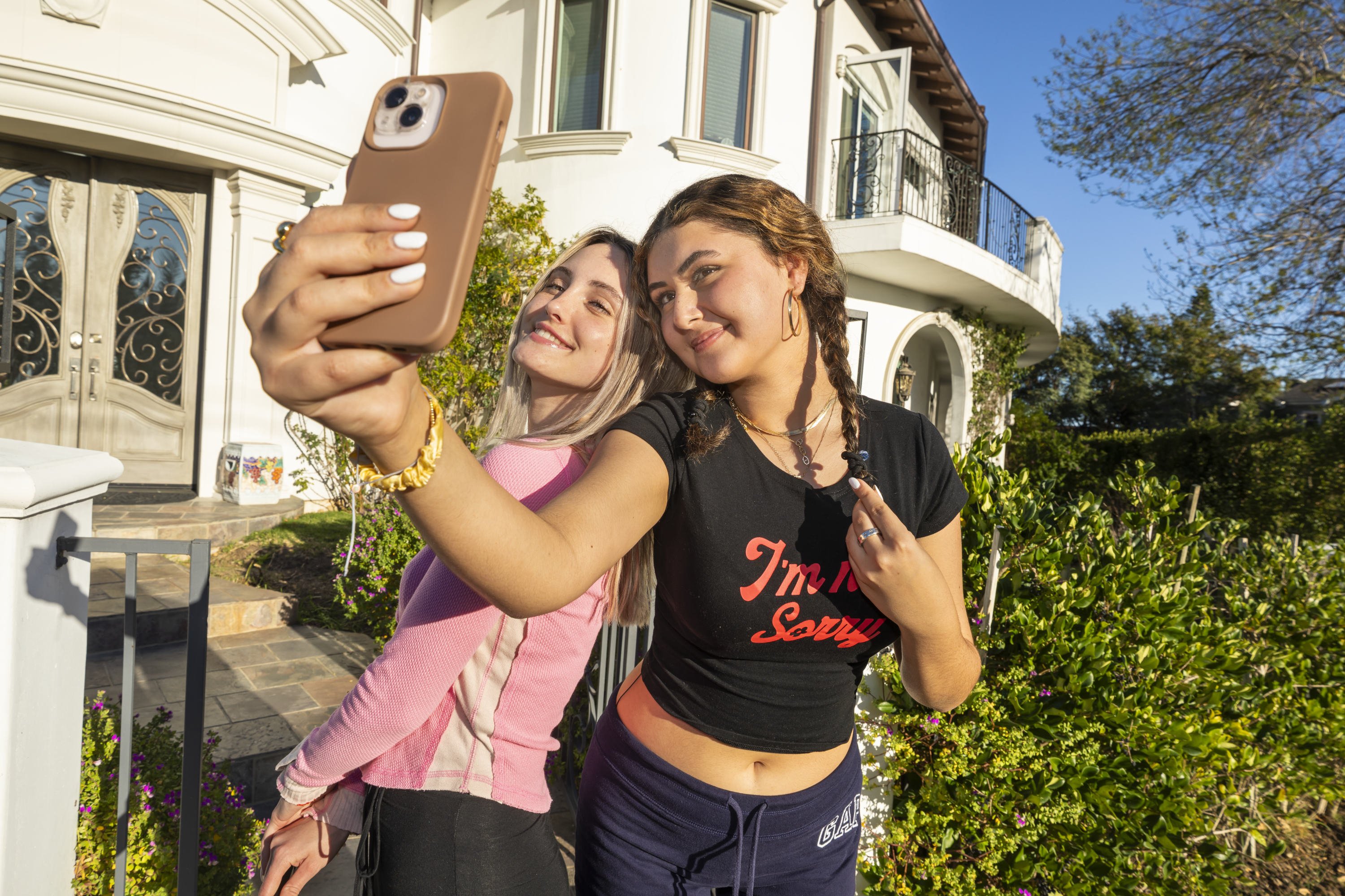 Zwei junge Frauen stehen vor einer weißen Villa dicht beieinander und machen ein Selfie; eine hält das Smartphone hoch, beide lächeln in die Kamera.